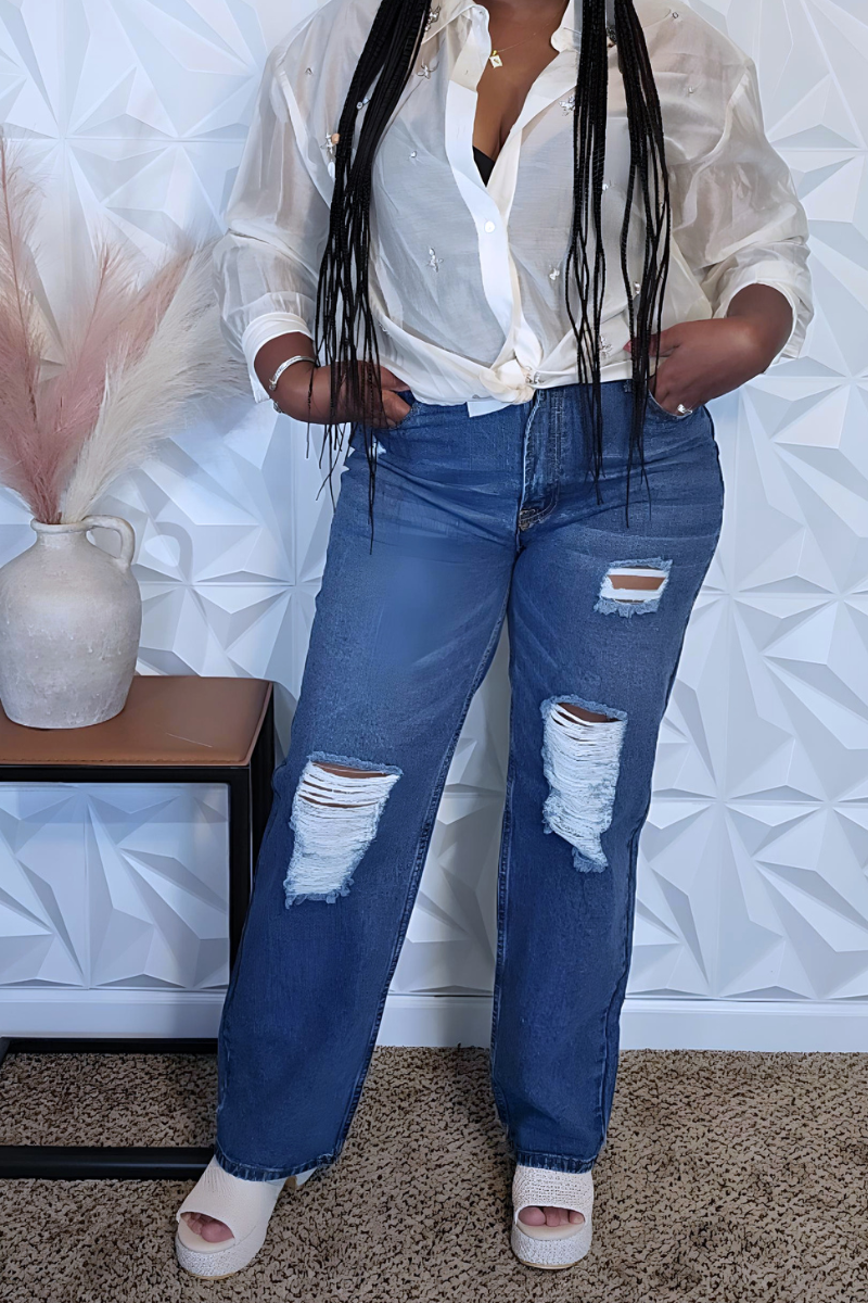 A person with long braided hair stands confidently against a white textured wall. They wear a green NY cap, a casually tied white shirt, distressed Daisy High Waist Jeans from I Am Lilou Bloom, and white open-toe heels. A table with a vase of decorative grass is beside them on the left.