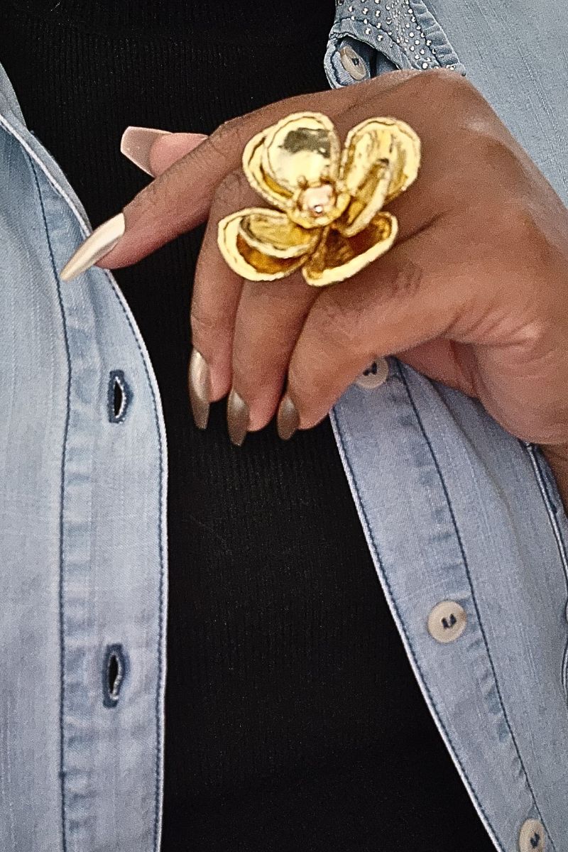 A person wearing a light denim button-up shirt, unbuttoned over a black shirt, holds their hand near their chest. They showcase long, metallic, pointed nails and wear a large Flower Petal Gold Ring by I Am Lilou Bloom on their middle finger.