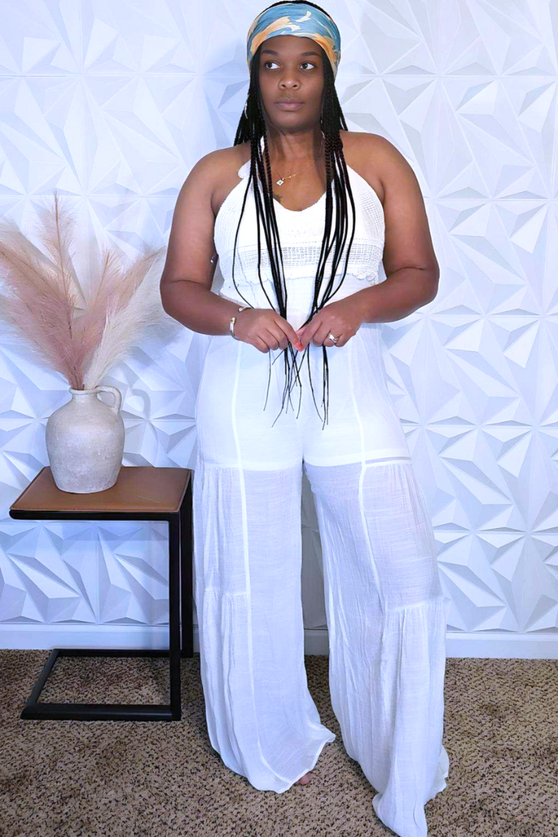 A woman with long, curly hair stands against a cream-colored wall wearing the Tulum Babe Linen Jumpsuit from Hyfve. She accessorizes with colorful bracelets and a layered necklace. In the background, small framed art pieces adorn the wall.