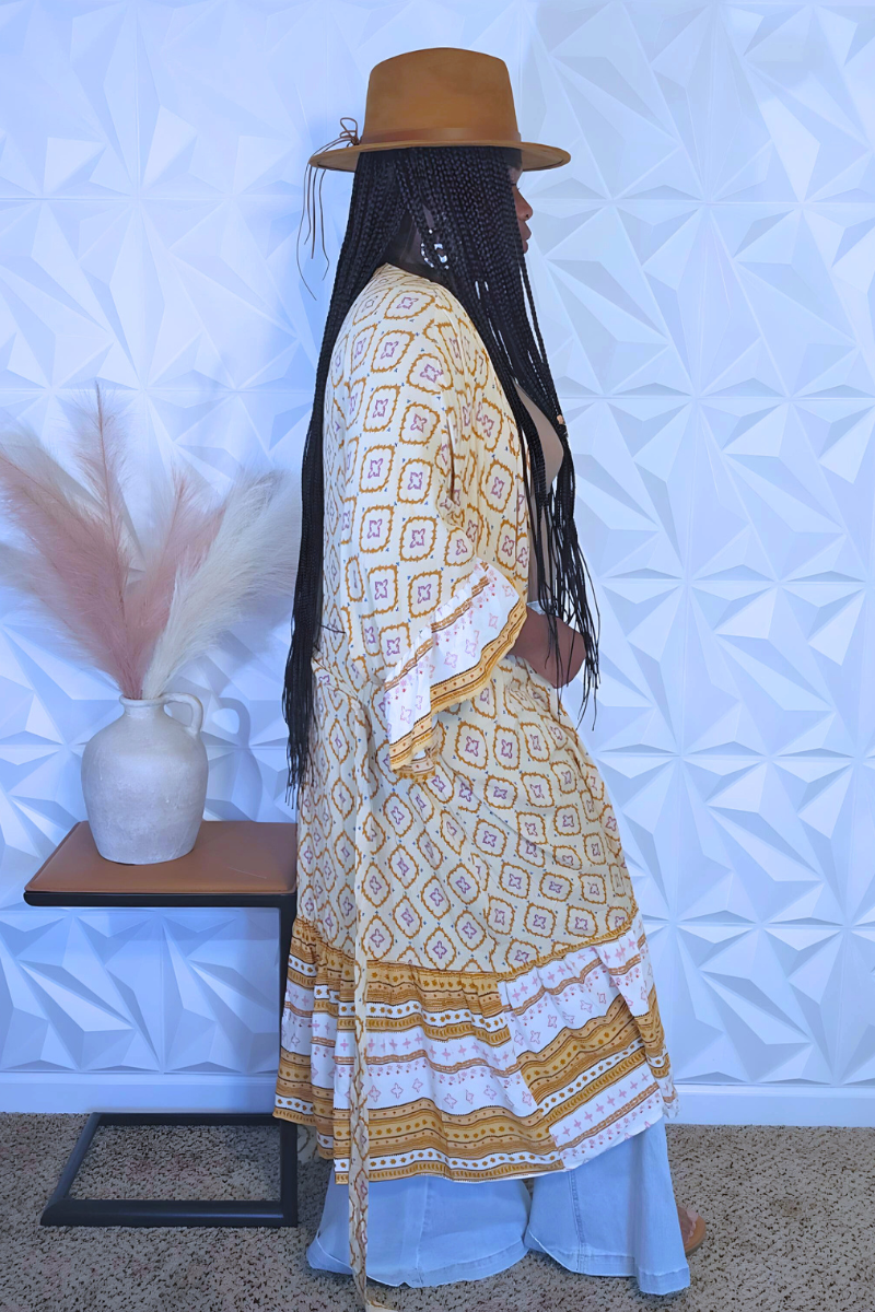 A person stands in front of a textured white wall wearing a brown hat, an I Am Lilou Bloom Women's 3/4 Ruffle Sleeve Self Tie Multi Print Kimono, wide-leg jeans, and holding a mustard yellow clutch. They have long braided hair and are standing beside a small table with a vase of pampas grass on it.