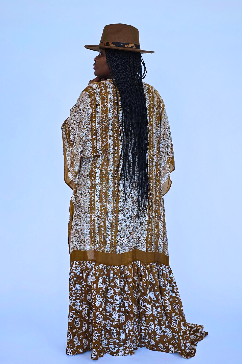 A person stands confidently against a textured white wall, wearing a vibrant orange head wrap and large earrings. They are draped in the I Am Lilou Bloom Flowy Paisley Boho Duster Kimono over a white top with light blue flared jeans. A table with a white vase of dried flowers is next to them, truly showcasing this versatile summer wardrobe must-have.