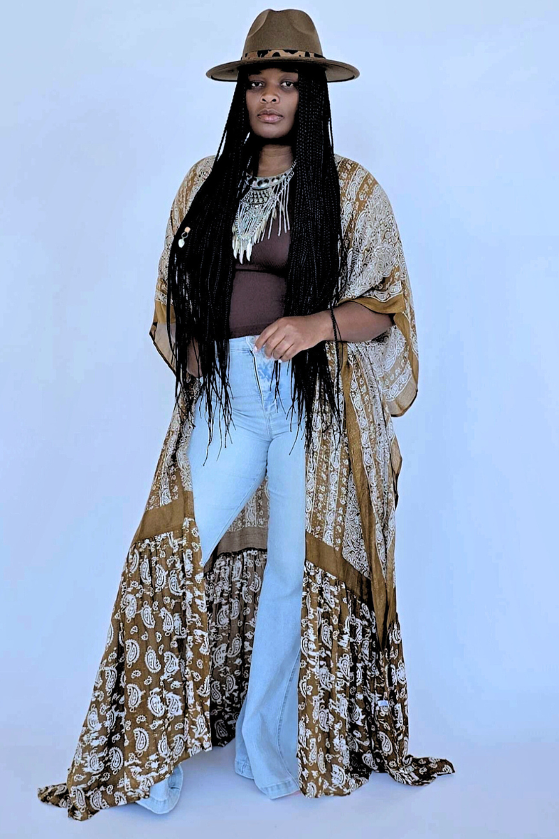 A person stands confidently against a textured white wall, wearing a vibrant orange head wrap and large earrings. They are draped in the I Am Lilou Bloom Flowy Paisley Boho Duster Kimono over a white top with light blue flared jeans. A table with a white vase of dried flowers is next to them, truly showcasing this versatile summer wardrobe must-have.