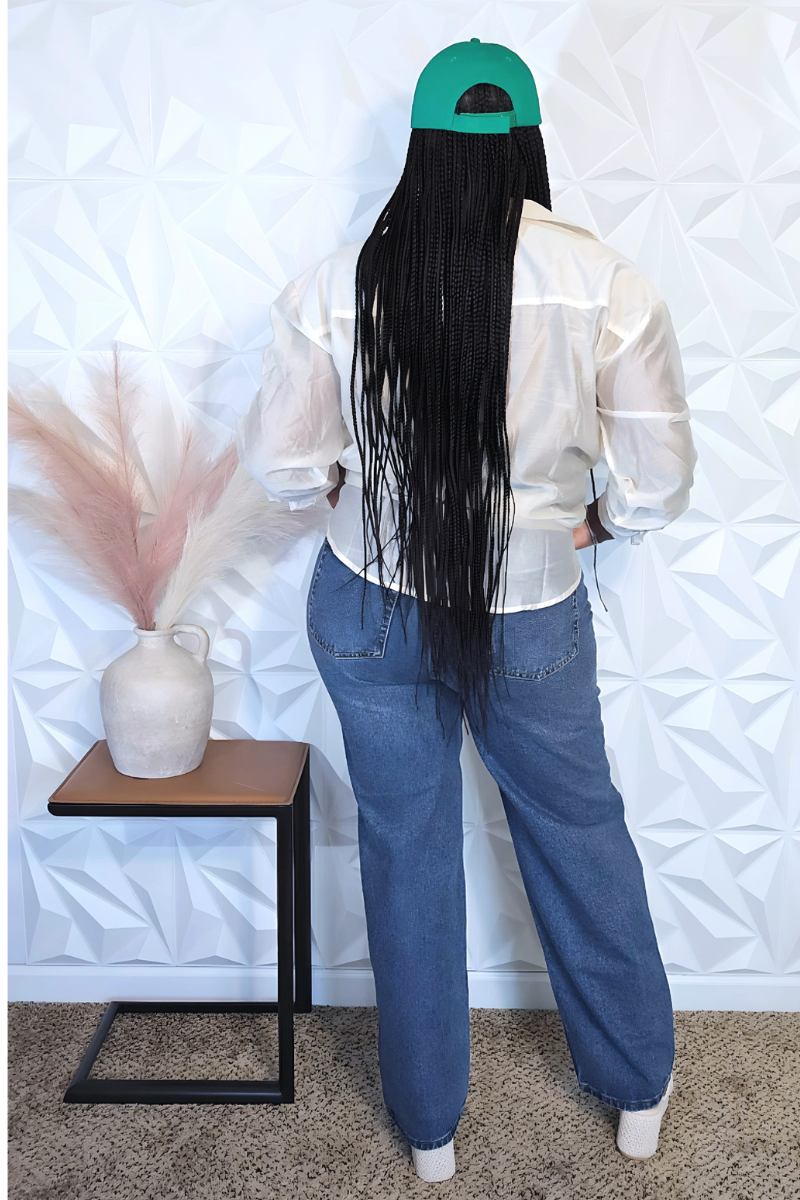 A person with long braided hair stands confidently against a white textured wall. They wear a green NY cap, a casually tied white shirt, distressed Daisy High Waist Jeans from I Am Lilou Bloom, and white open-toe heels. A table with a vase of decorative grass is beside them on the left.