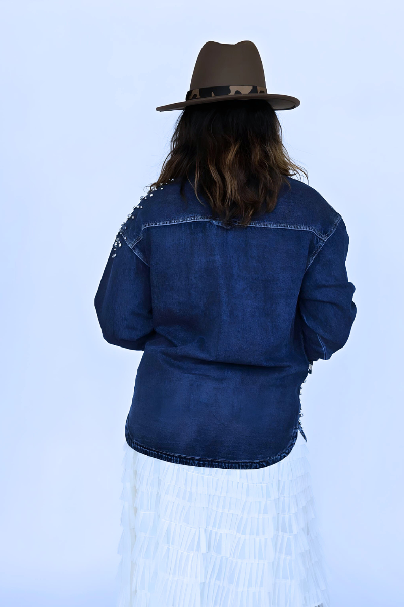A person is wearing the "Embellished Denim Shirt" by I Am Lilou Bloom, a relaxed fit, blue cotton denim overshirt adorned with delicate pearl and crystal stud embellishments. They are accessorized with gold bracelets on their left wrist and a gold necklace. The shirt is partially unbuttoned, revealing a bit of skin underneath. Their nails are painted white.