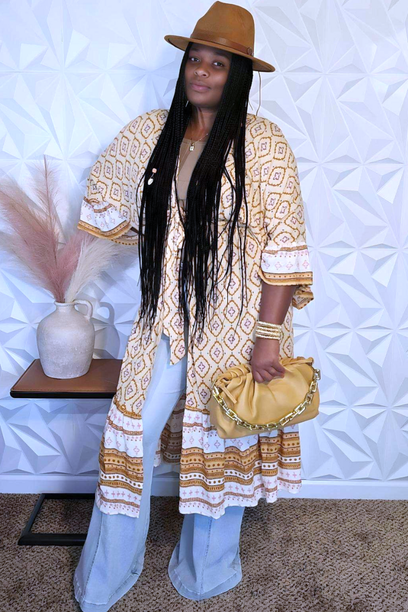 A person stands in front of a textured white wall wearing a brown hat, an I Am Lilou Bloom Women's 3/4 Ruffle Sleeve Self Tie Multi Print Kimono, wide-leg jeans, and holding a mustard yellow clutch. They have long braided hair and are standing beside a small table with a vase of pampas grass on it.
