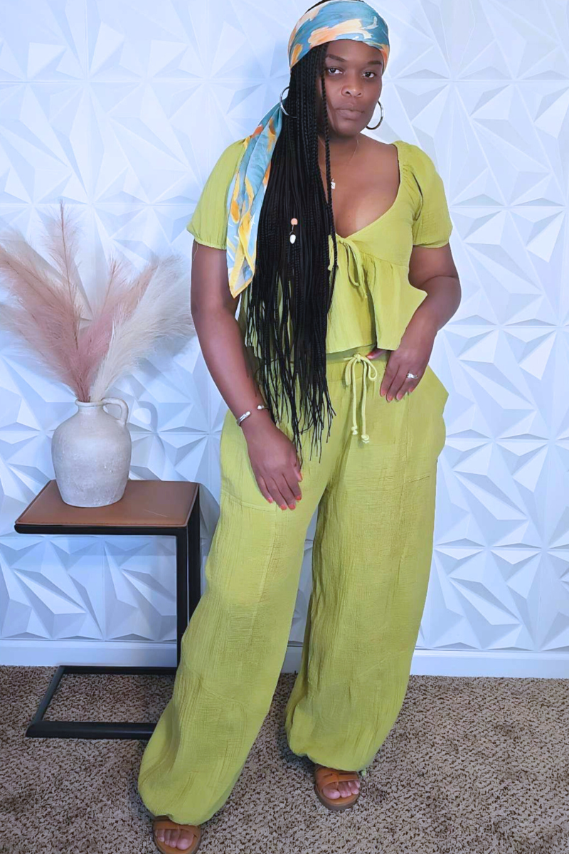 A woman stands indoors wearing the Bali Babe Green Set from Hyfve, which includes a lightweight crop top and matching wide-leg pants. With long braided hair and a colorful headscarf, she poses in front of a small table adorned with a decorative vase and dried pampas grass.
