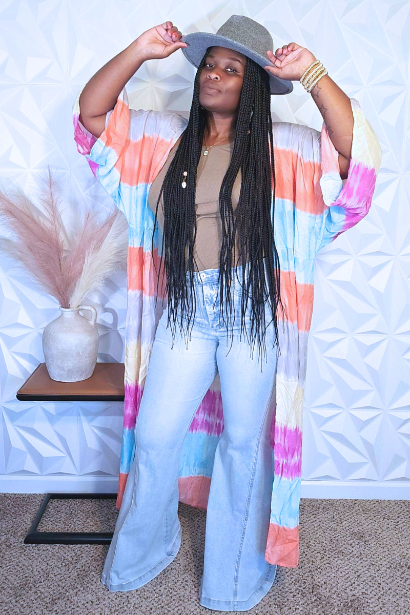 A person with long braided hair stands sideways, wearing a colorful dress from I Am Lilou Bloom that resembles the Cotton Candy Kimono, paired with a gray hat. They are near a small table with a beige vase filled with dried pampas grass. The background features a geometric white wall pattern.