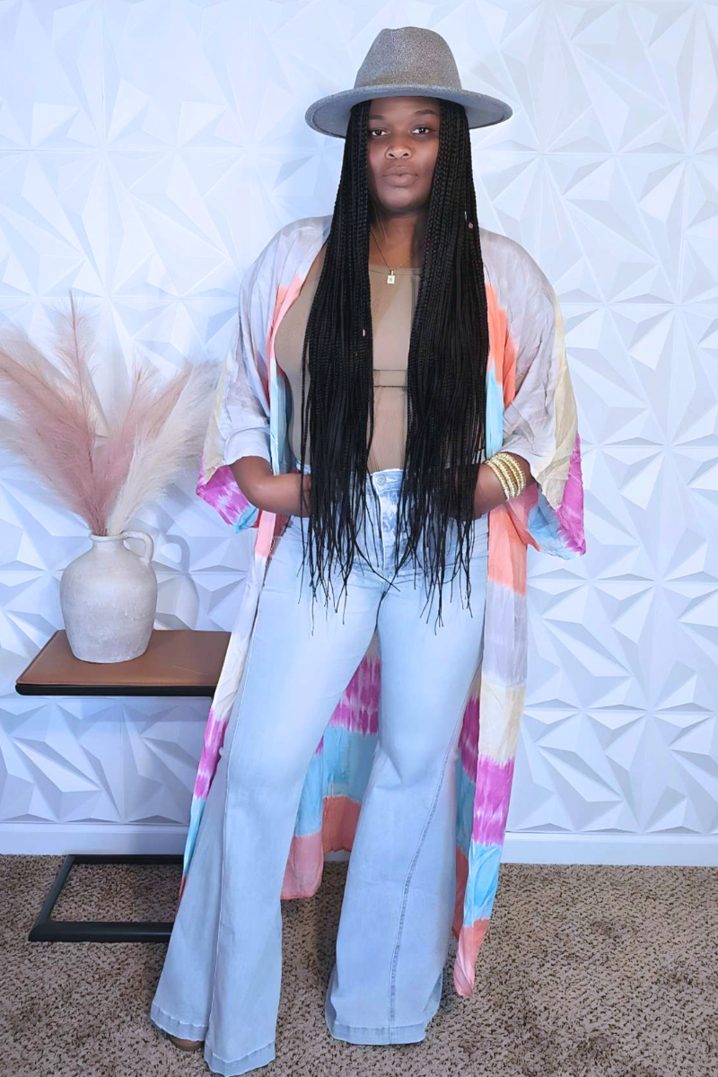 A person with long braided hair stands sideways, wearing a colorful dress from I Am Lilou Bloom that resembles the Cotton Candy Kimono, paired with a gray hat. They are near a small table with a beige vase filled with dried pampas grass. The background features a geometric white wall pattern.