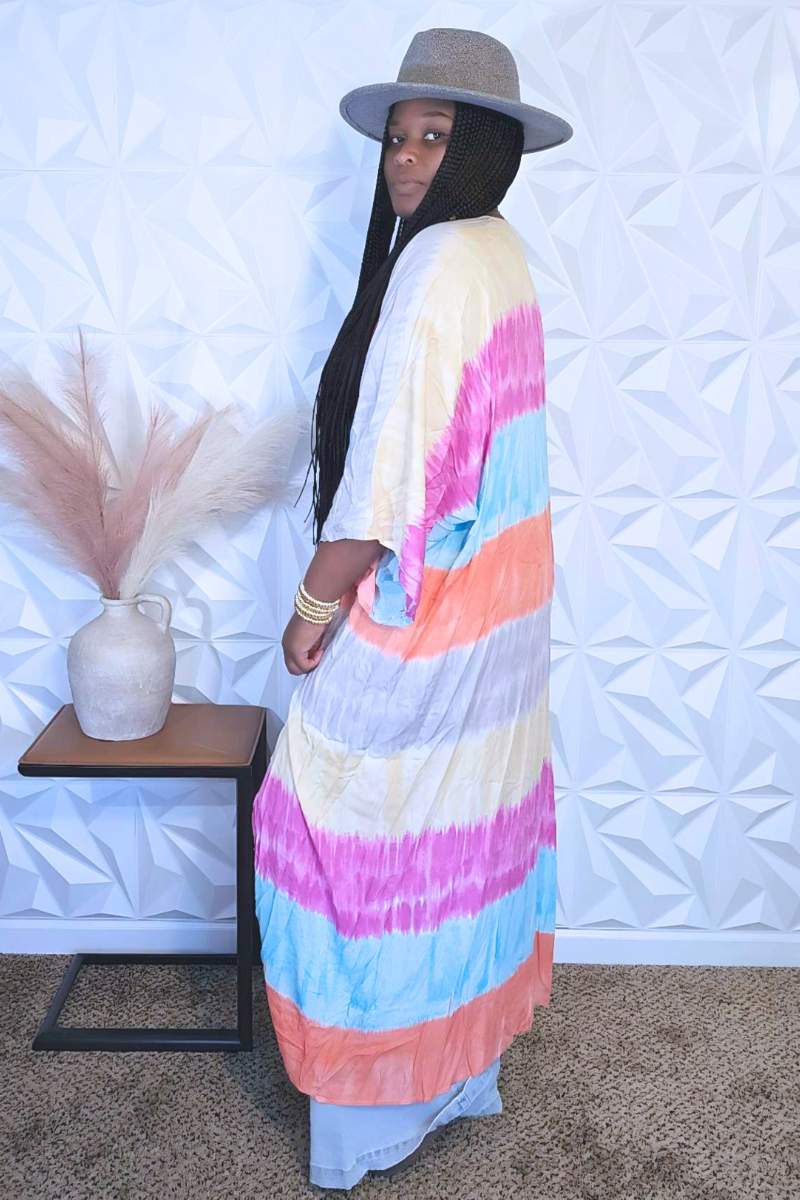 A person with long braided hair stands sideways, wearing a colorful dress from I Am Lilou Bloom that resembles the Cotton Candy Kimono, paired with a gray hat. They are near a small table with a beige vase filled with dried pampas grass. The background features a geometric white wall pattern.