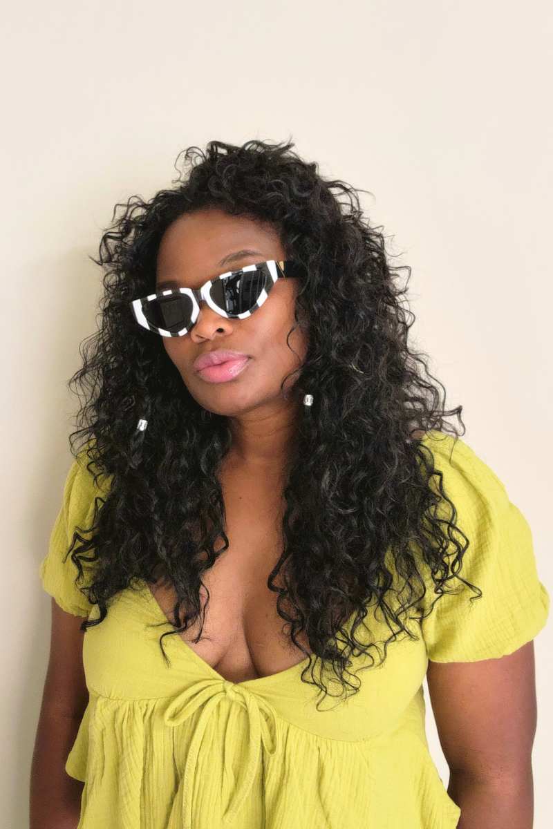 A person with long, curly black hair wearing I Am Lilou Bloom's Shay Elegant Sunglasses and a light green, short-sleeved top stands against a plain, light-colored background. The person's expression is neutral.